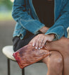 A woman sitting on a bench holding a Bed Stu Cadence clutch.