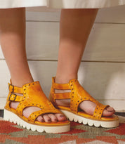 A woman wearing Bed Stu Camden sandals standing on a rug.