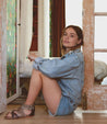 A woman sitting on the floor in a Bed Stu Capriana denim jacket and shorts.