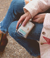 A woman sitting on a bench holding a Bed Stu wallet named Carrie.