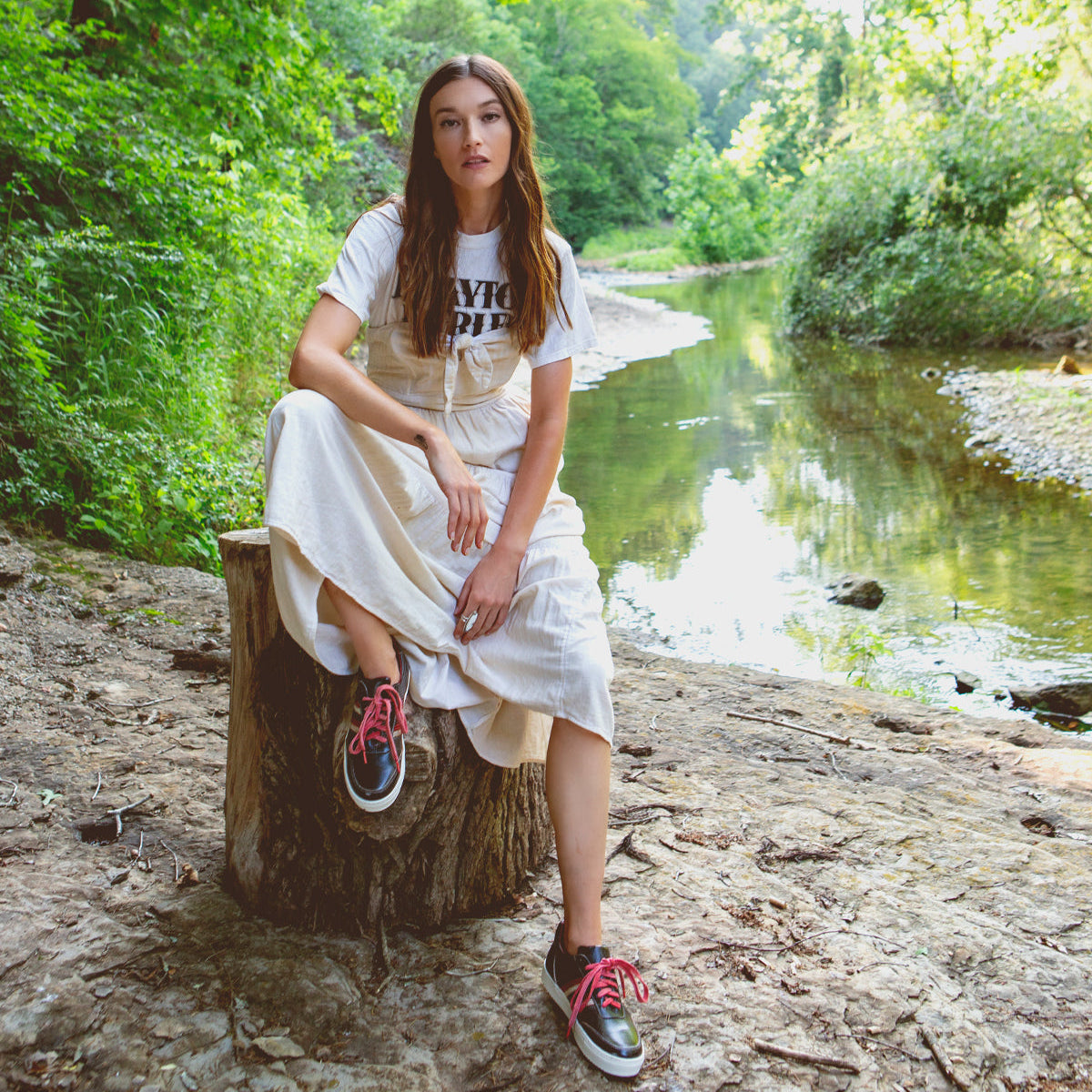 A woman is sitting on a Carrington stump next to a river by Bed Stu.