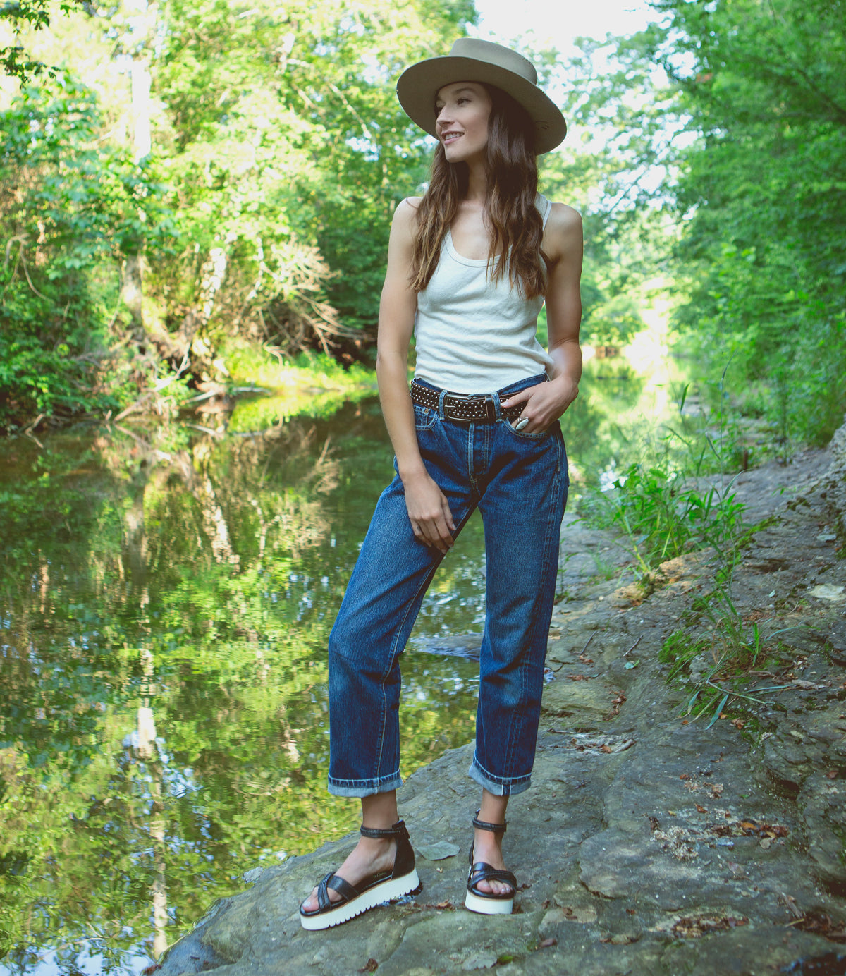 A woman wearing jeans and a Caroll sandals by Bed Stu standing by a river.