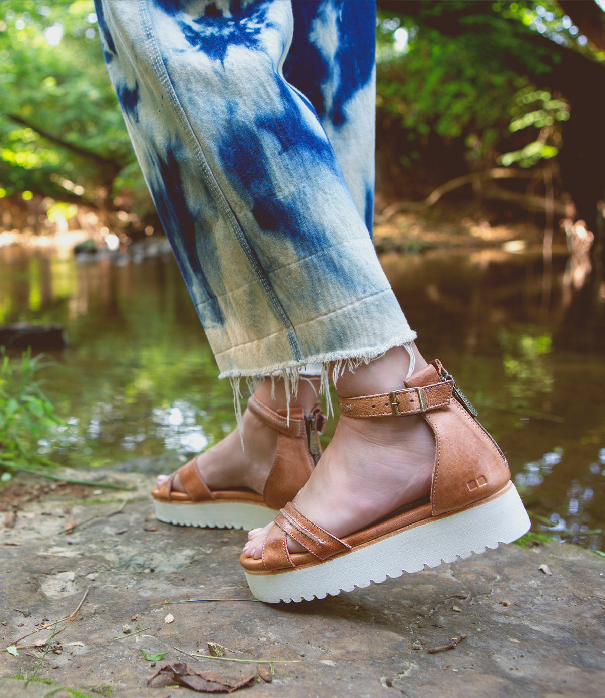 A person wearing Caroll Sandals by Bed Stu sandals standing on a rock next to a river.