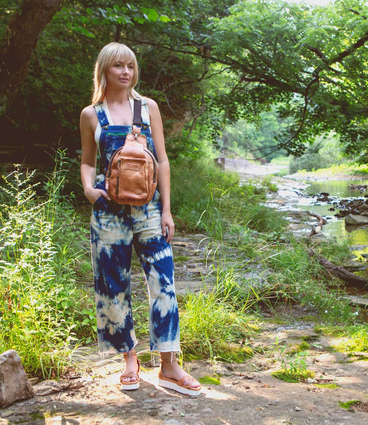 A woman in a jumpsuit wearing Caroll sandals by Bed Stu, standing near a river.