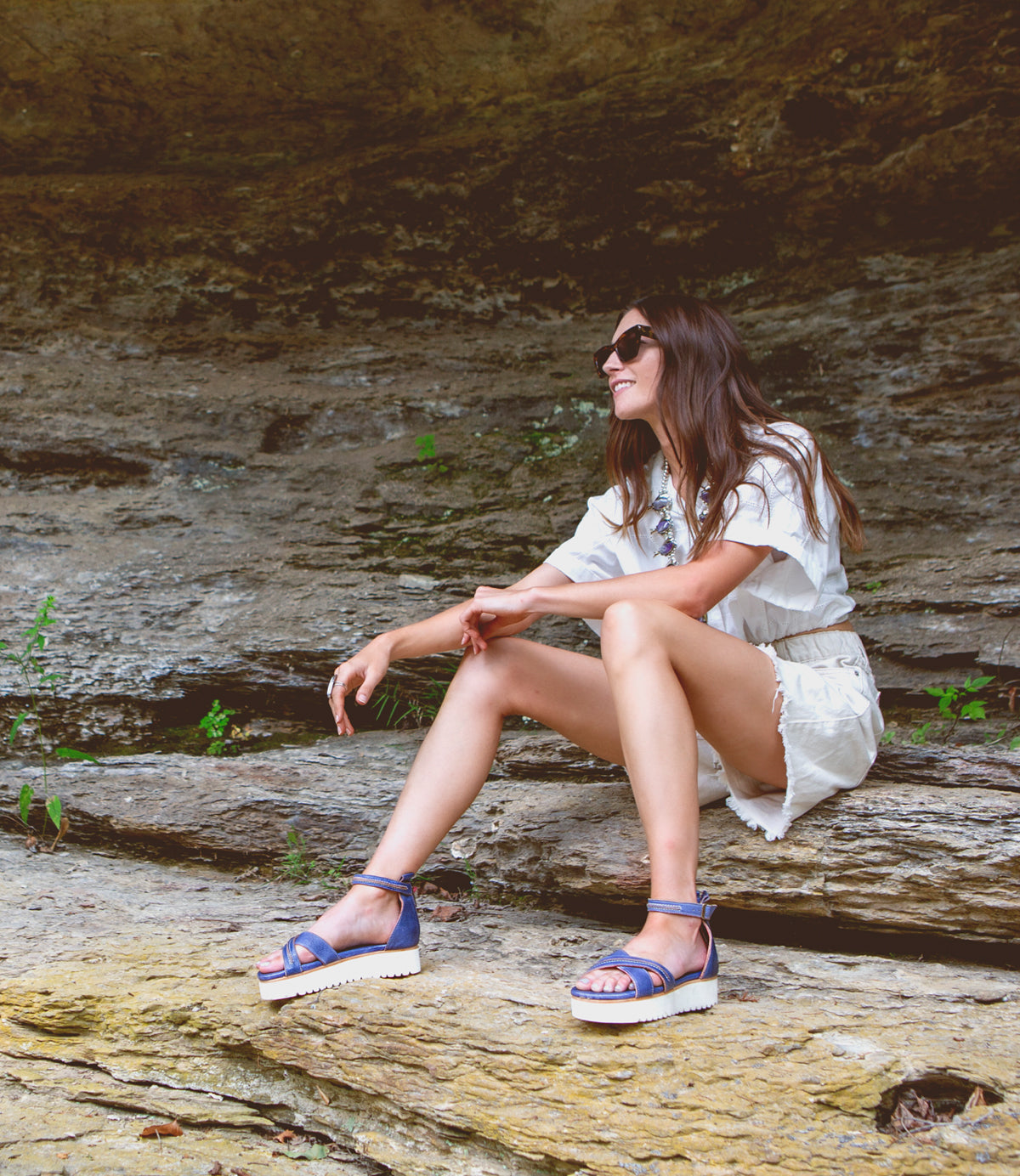 A woman is sitting on a rock and wearing Bed Stu sandals, called Carroll.