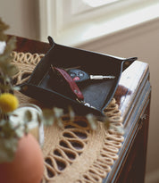 A black Expanse Catchall Tray by Bed Stu holding a set of car keys, a multi-tool knife, and some loose change sits on a wooden surface next to a window.
