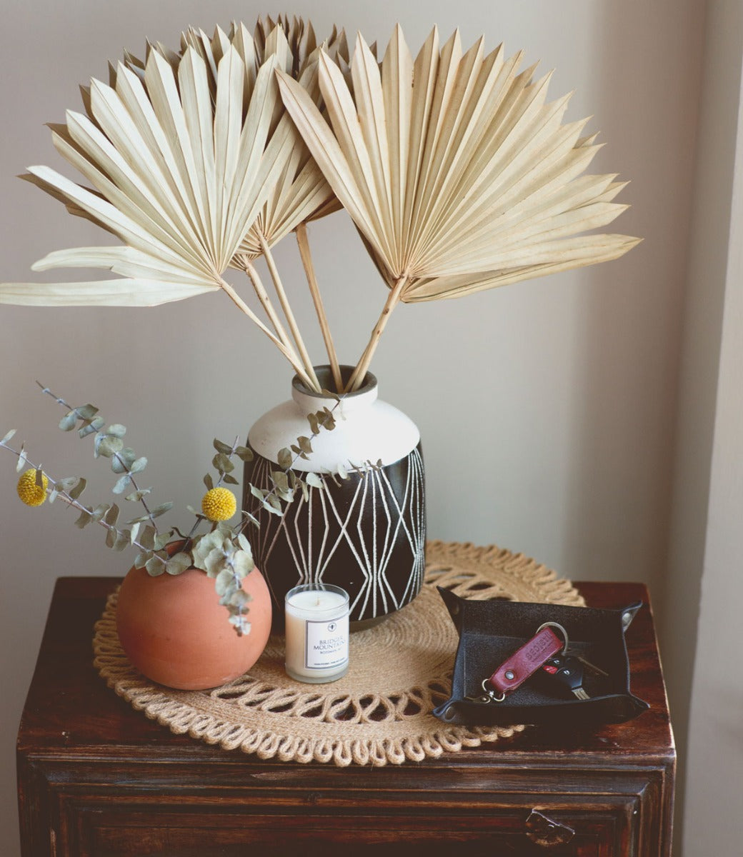 A Expanse vase with palm leaves and a candle on a table by Bed Stu.