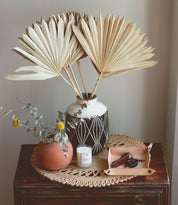 A decorative setup on a wooden surface includes a vase with dried palm leaves, a smaller vase with eucalyptus, a candle, and an Expanse Catchall Tray by Bed Stu holding keys, loose change, and a pocketknife, all placed on a doily underneath.