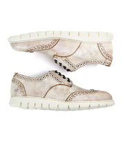 The Bed Stu Cayuga II is a pair of light beige, lightweight leather wingtip derby shoes featuring stylish brogue details and white rubber soles. Positioned side by side, with one sole facing up and the other down, these contemporary casual male shoes offer both elegance and comfort.