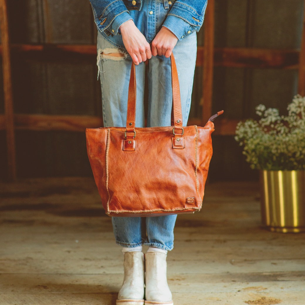A woman holding a Celindra LTC by Bed Stu brown leather tote bag.