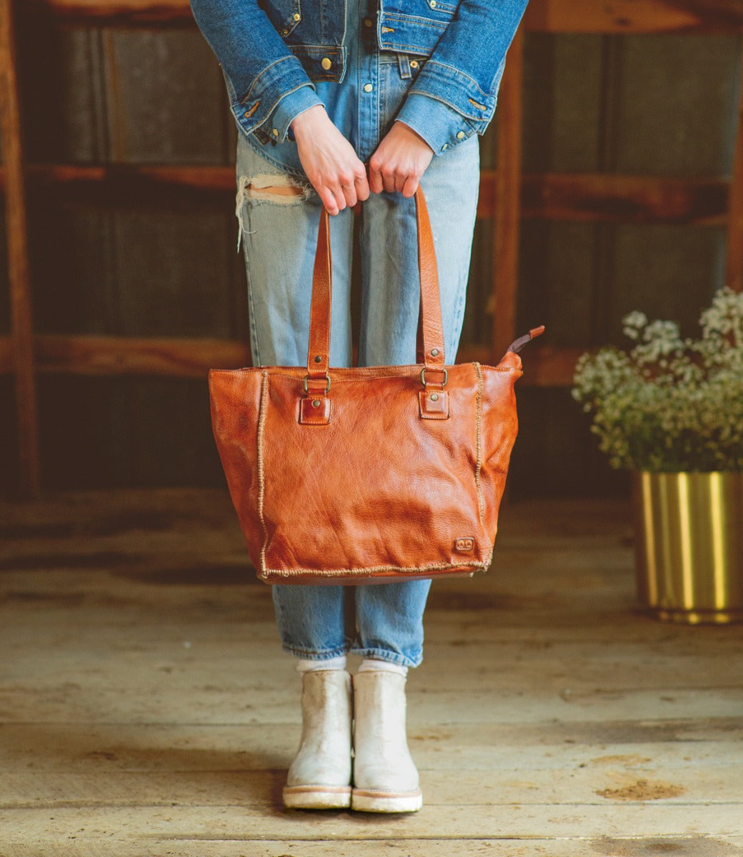 A woman holding a Celindra LTC by Bed Stu brown leather tote bag.
