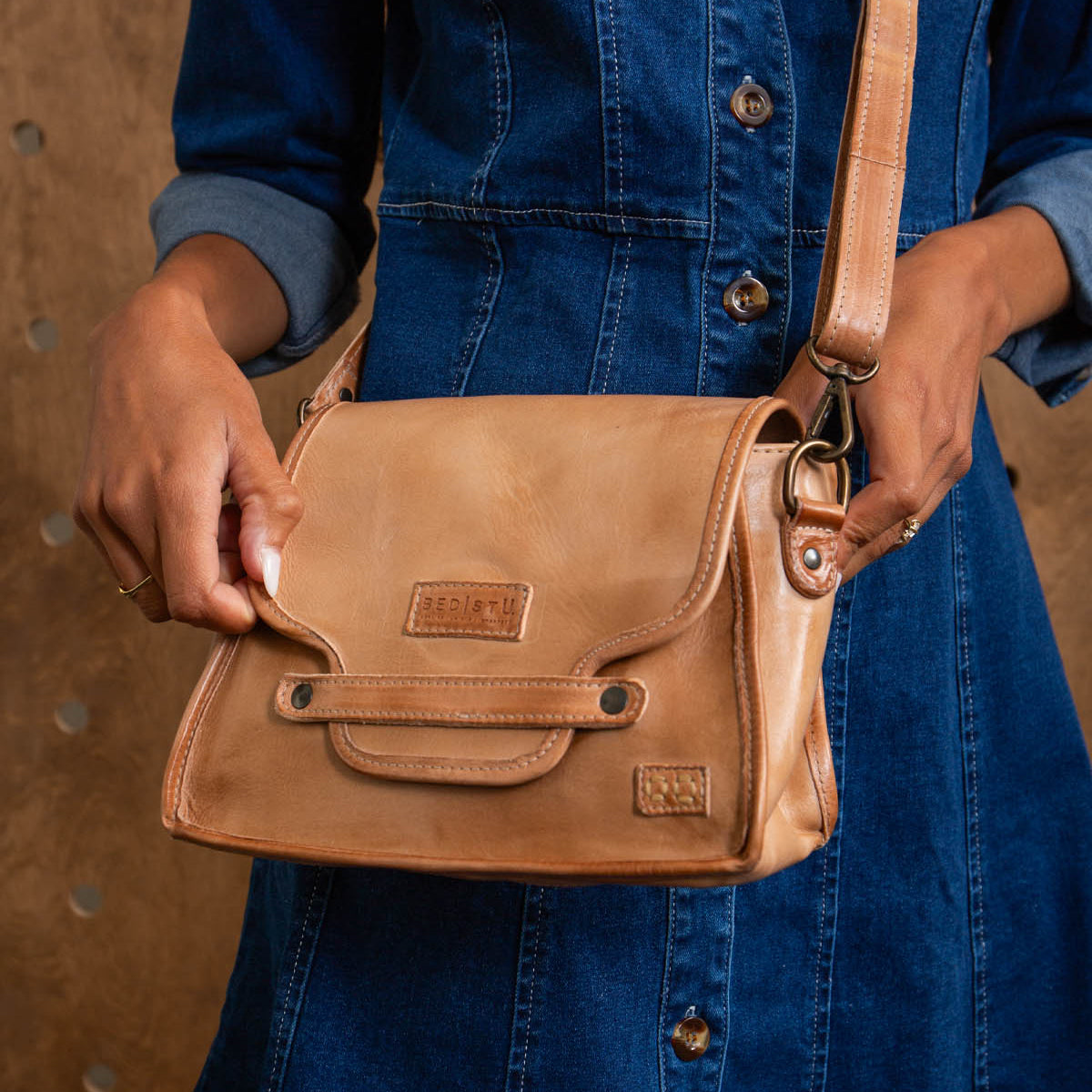 A person in a blue denim dress holds and opens the Bed Stu Ceremonious light brown leather flap-top bag, featuring a magnetic closure and an embossed logo on the front.