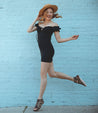 A woman in a black off the shoulder Bed Stu dress jumping against a brick wall.