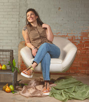 A woman sits on a white chair, smiling in her checkered top and jeans. Pears rest on the fabric-draped floor, set against a brick backdrop. Her relaxed style is enhanced by the metal grommet detail of her Clancy shoes by Bed Stu.
