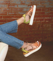 Feet in Bed Stu's Clancy sandals, metallic copper and green with cushioned insoles and white platform soles, rest on a beige chair. Denim jeans against a brick wall complete the scene.