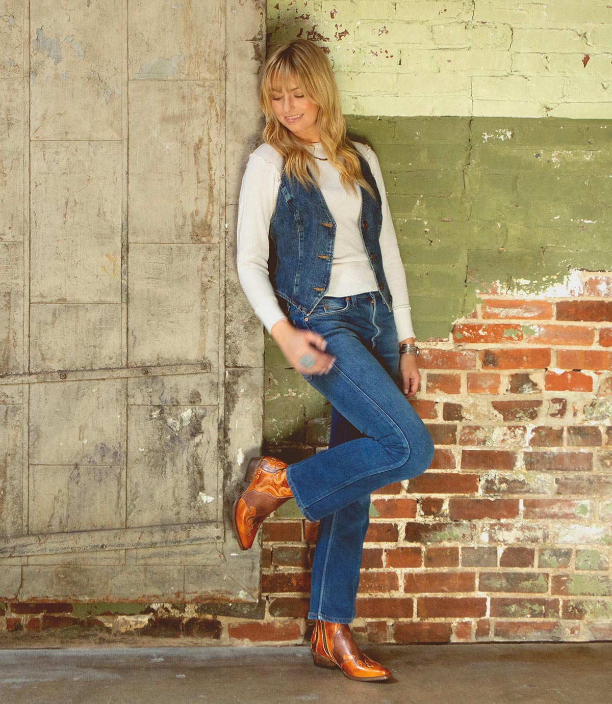 A woman in jeans and leather boots leaning against a rustic brick wall, showcasing her Bed Stu Deuce western style.