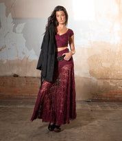 A woman indoors poses against a textured wall, wearing the Bed Stu Dreamweaver burgundy lace two-piece, styled with a vintage boho belt and holding a black jacket over her shoulder.