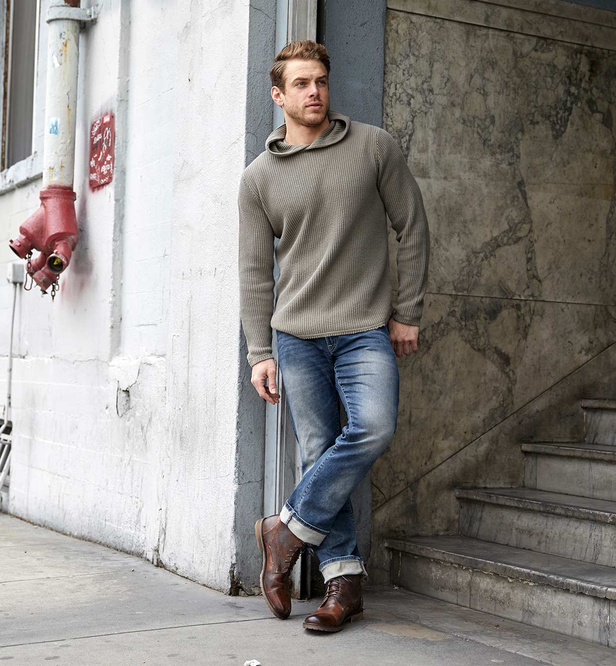 A man in Bed Stu jeans and a Bed Stu hoodie leaning against a wall.