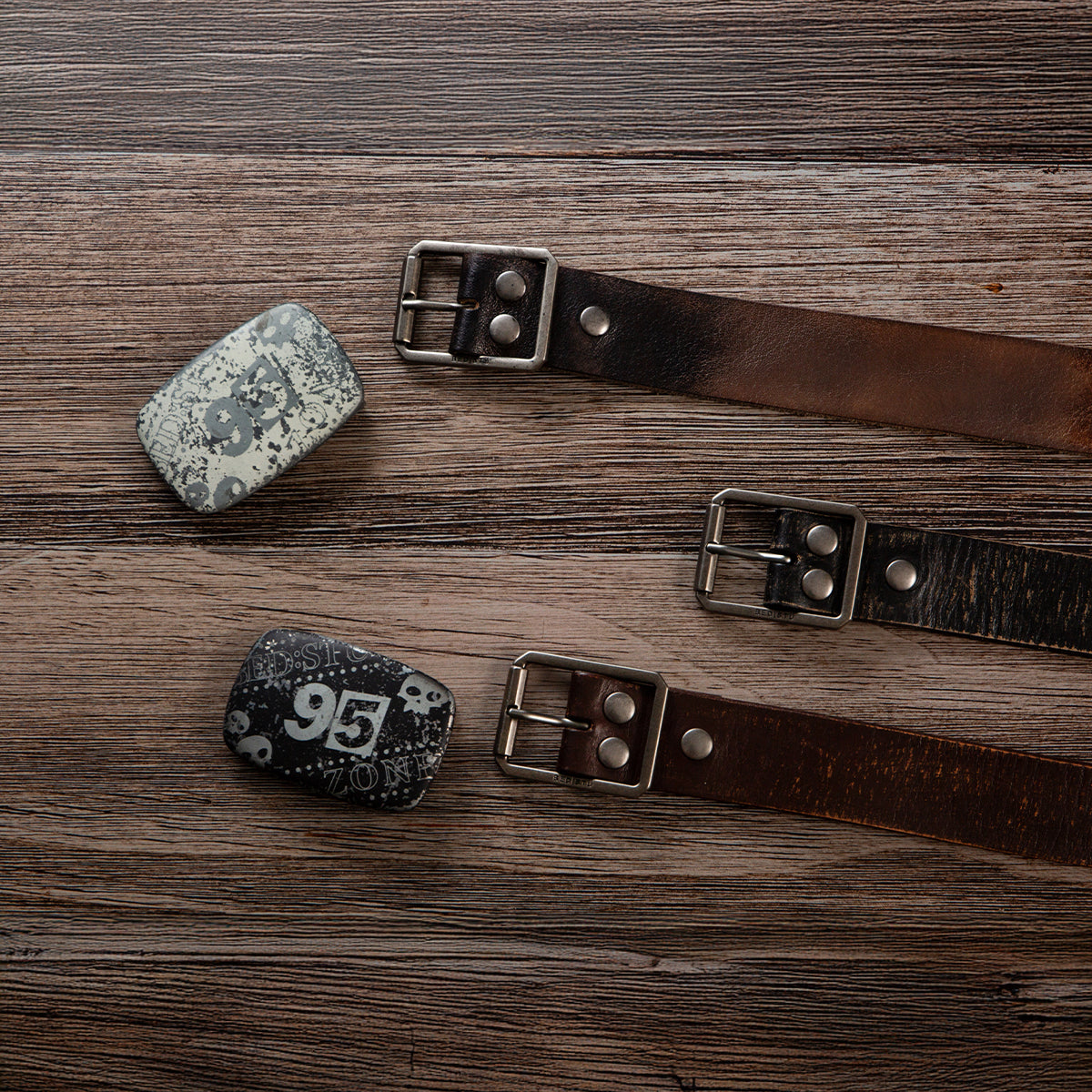 The Bed|Stü Drifter Belt Buckle Bundle includes two premium leather belts with metal buckles, plus two vintage-style belt buckles numbered "95" and "96," all displayed on a wooden surface.