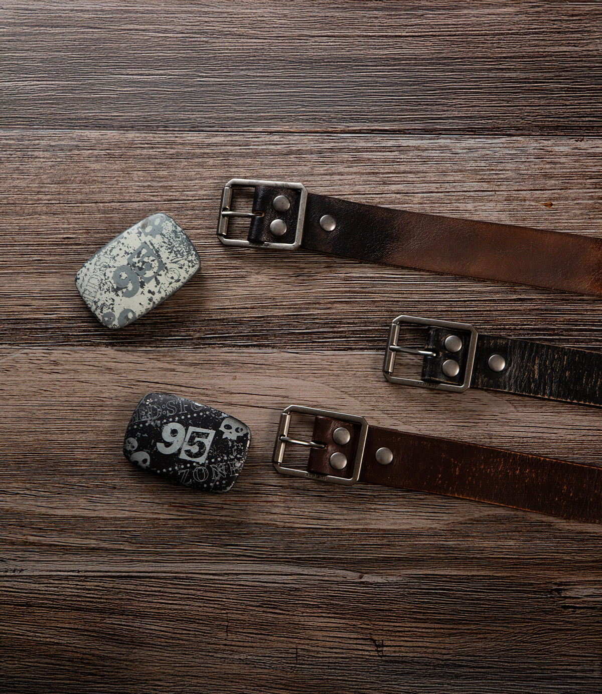 The Bed|Stü Drifter Belt Buckle Bundle includes two premium leather belts with metal buckles, plus two vintage-style belt buckles numbered "95" and "96," all displayed on a wooden surface.