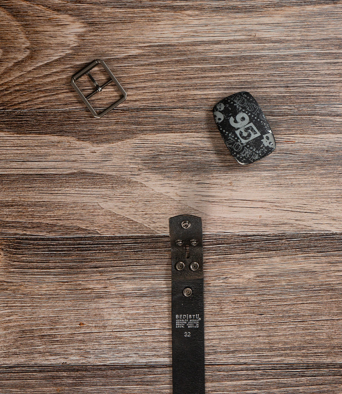 The Bed|Stü Drifter Belt Buckle Bundle, featuring premium leather, a metal buckle, and a graphic belt end, is disassembled and laid out on a wooden surface.