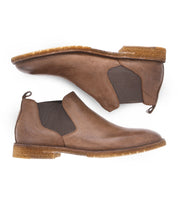 The Elude by Bed Stu are brown leather Chelsea boots with elastic side panels and tan rubber soles, photographed side-by-side, sole to sole. These men's boots showcase a timeless design, expertly handcrafted in Italy for a perfect blend of style and elegance.