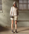 A woman in a beige Encase sweater and shorts standing in a warehouse, by the brand Bed Stu.