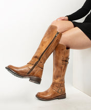 A person in a black dress wears Bed Stu's Essence tan knee-high riding boots with side zippers and buckles, made from hand-finished leather, seated against a white background.