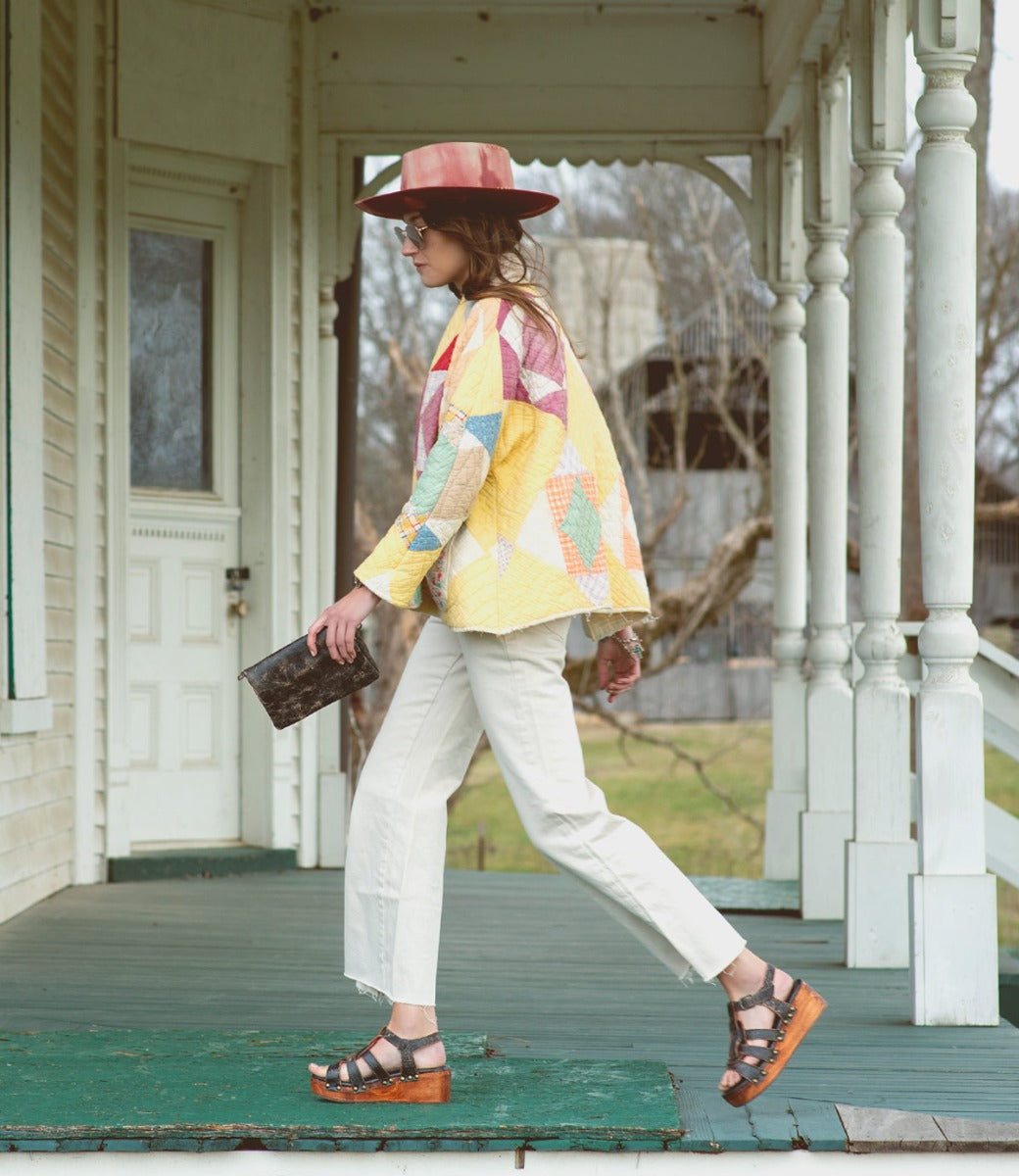 A woman in a yellow jacket and white pants walking on the porch of a Bed Stu distressed black Fabiola house.