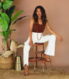 A woman sitting on a stool in white pants and a brown top, wearing Fabiola by Bed Stu.
