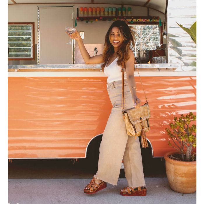 Woman standing in front of camper wearing a white shirt, tan pants, tan bag, and tan Fabiola sandals.