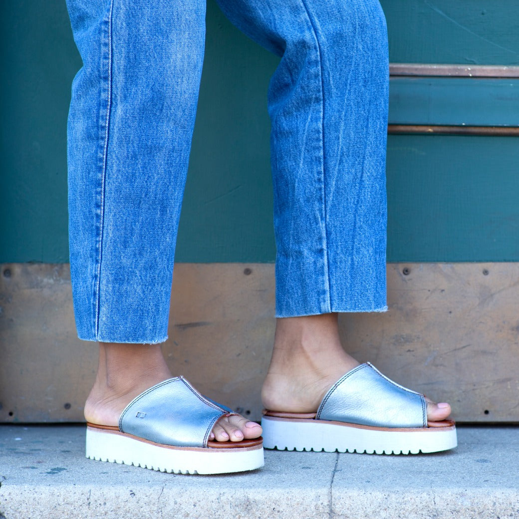A woman wearing a pair of Bed Stu Fairlee II silver slide sandals.