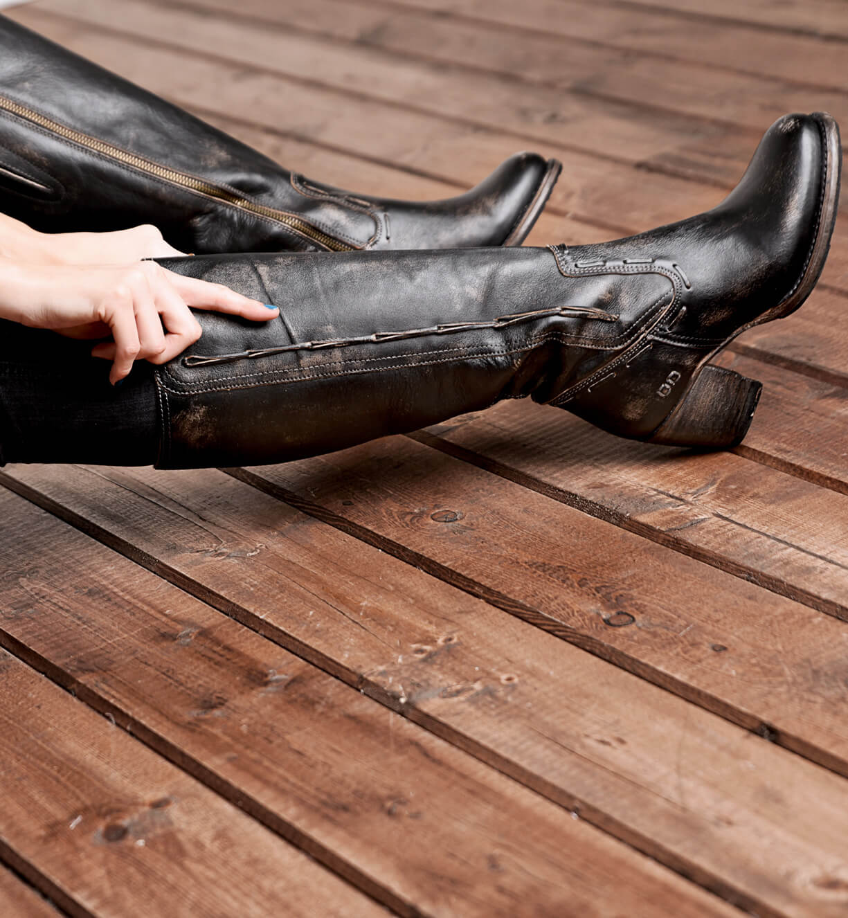 A woman wearing black Bed Stu boots sitting on a wooden floor.