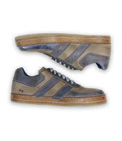 The Firepower sneakers by Bed Stu are men's shoes featuring gray and beige tones, two dark leather stripes on the sides, white laces, and a brown vulcanized sole.