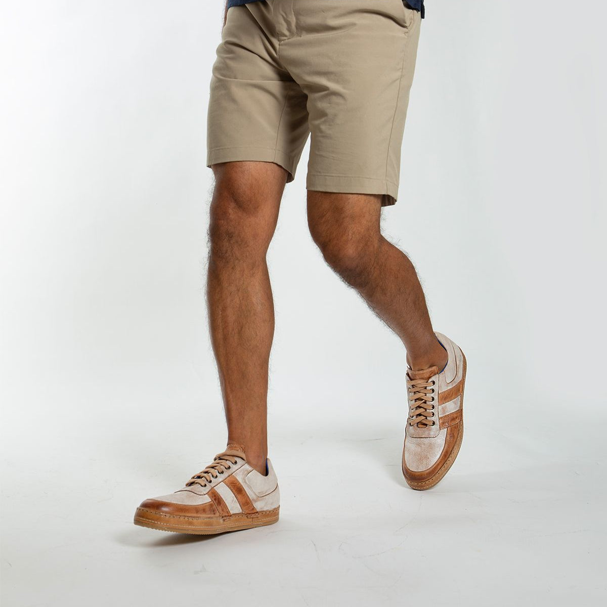 Wearing khaki shorts and a navy shirt, a person walks in Bed Stu's Firepower sneakers, featuring brown and white tones with leather stripe details. A plain white background highlights the vulcanized style of their footwear.