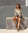 A woman sitting on a concrete wall wearing a Fontella gold jacket and shorts from Bed Stu.