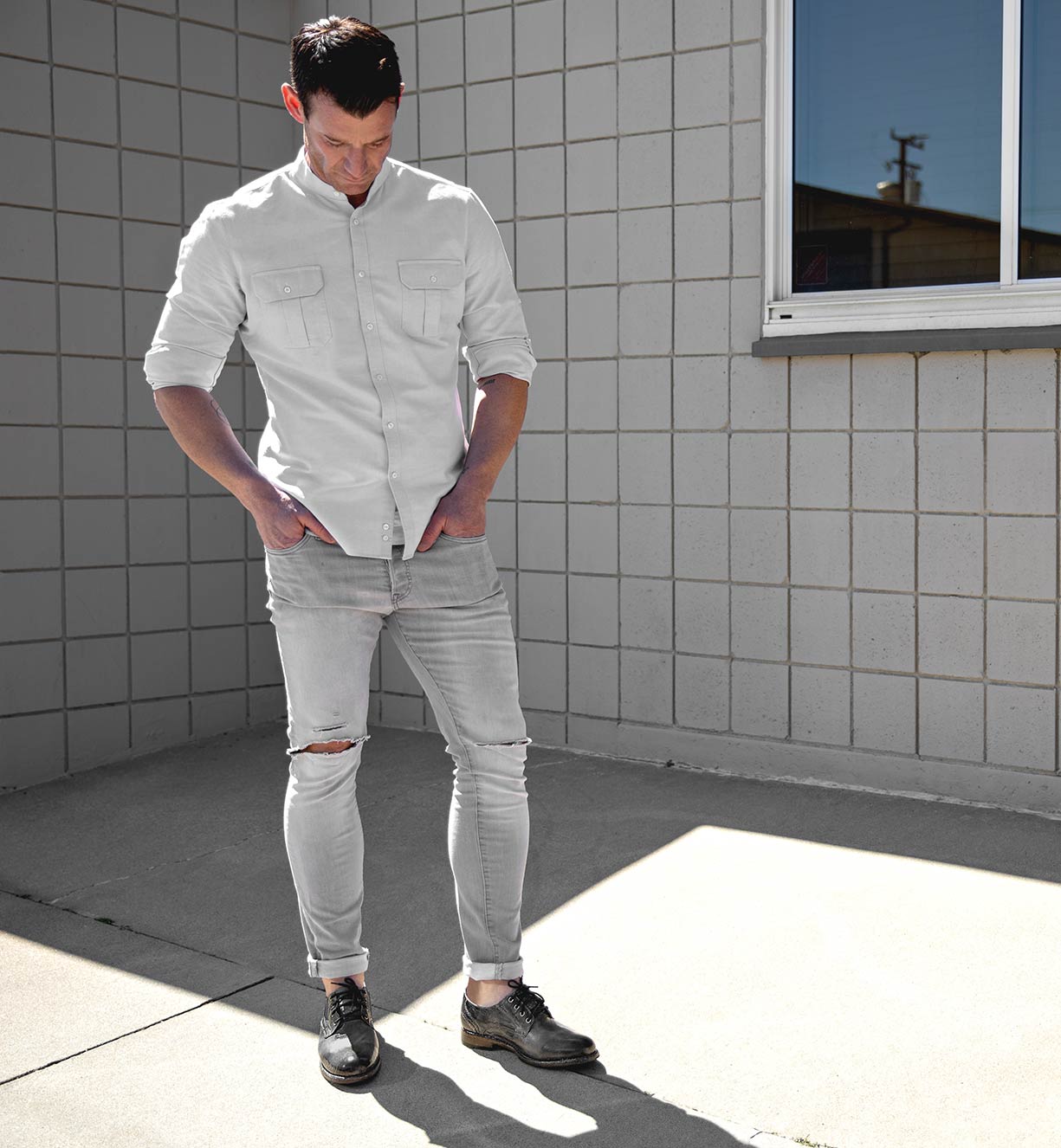 A man standing in front of a building wearing Bed Stu ripped jeans and a Bed Stu white shirt.