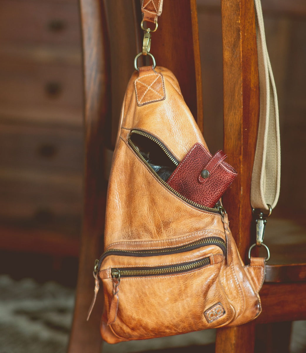 A brown leather Behold sling bag hanging on a chair from the Bed Stu brand.