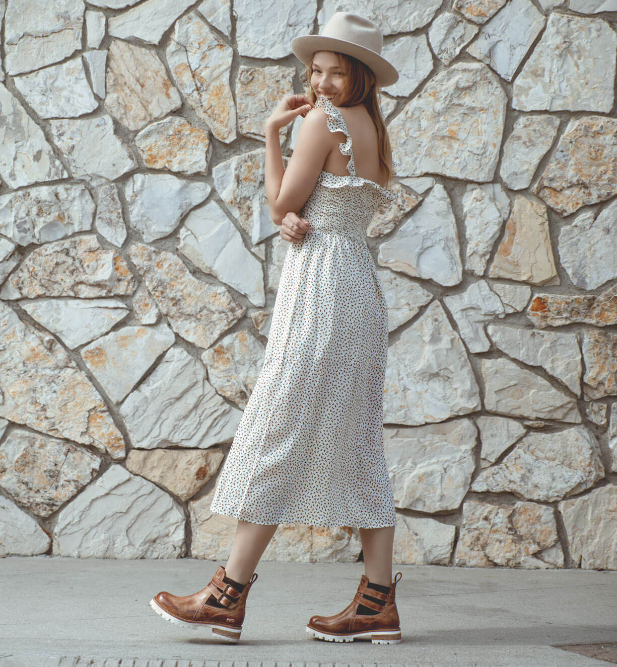 A woman wearing Ginger by Bed Stu, a hat and a dress standing in front of a stone wall.