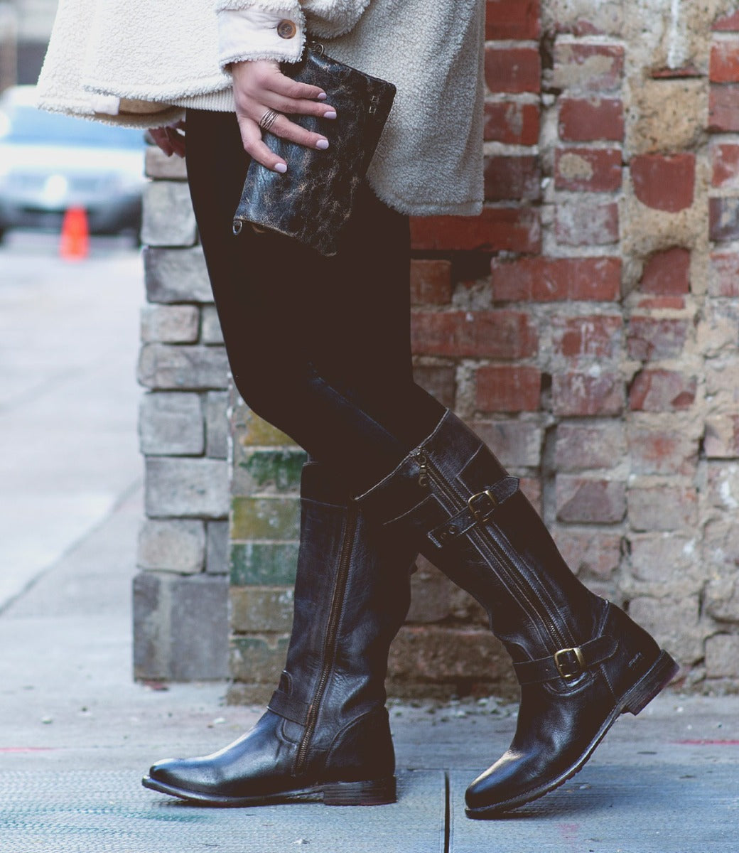 A woman in Gogo Lug Wide Calf boots by Bed Stu and a white sweater leaning against a brick wall.