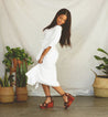 A woman in a white dress and brown Grettell sandals, posing in front of potted plants.