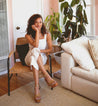 A woman sitting on a chair in a living room while wearing white shirt, white pants, and Grettell sandals..