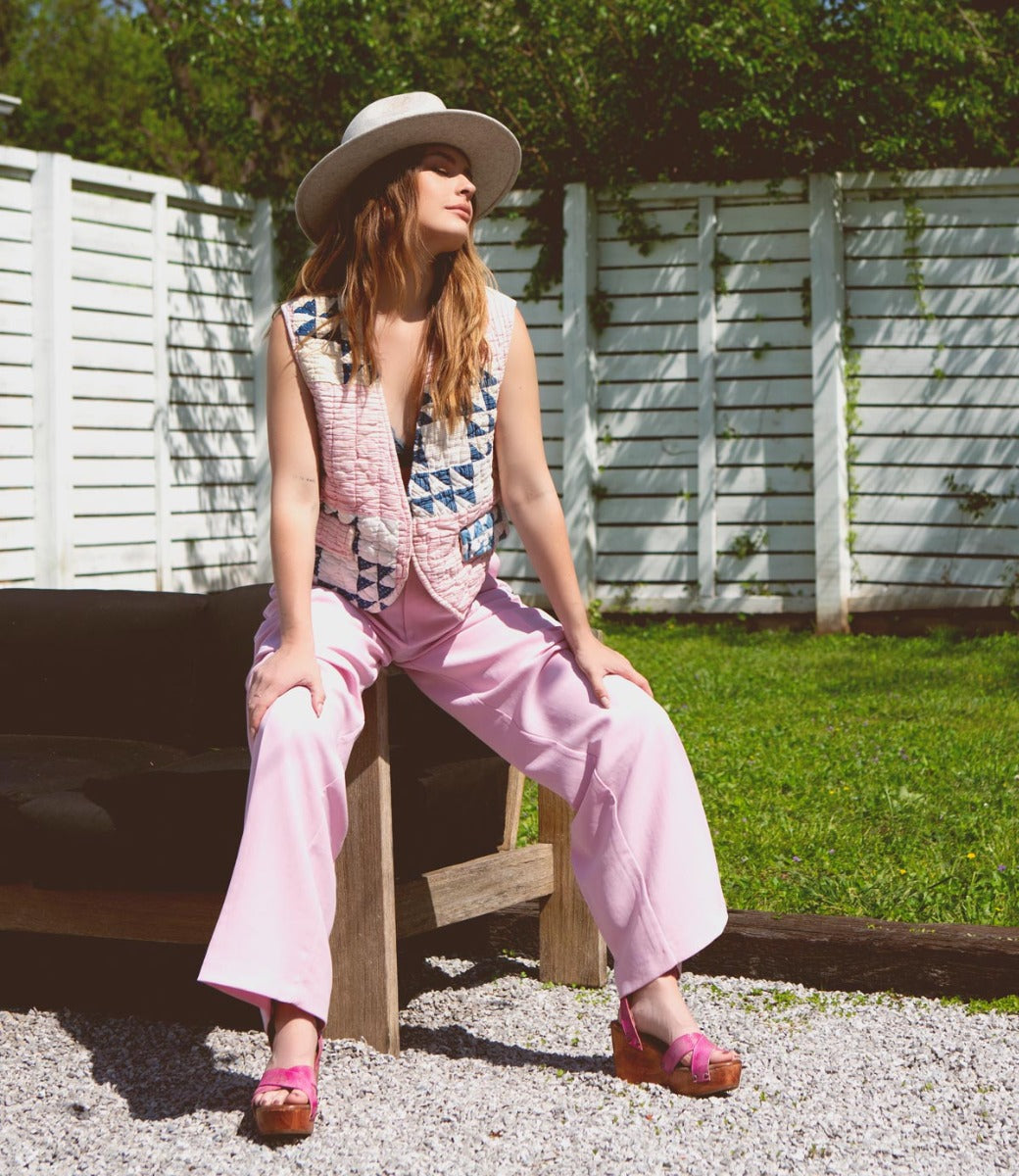 A woman wearing pint pants, pink vest, a hat and pink Grettell sandals sitting on a bench.