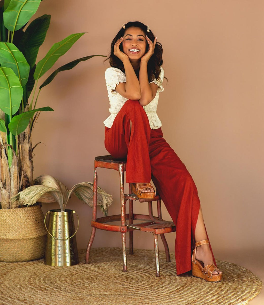 A woman sitting on a stooll wearing a white and yellow shirt, red pants and tan Grettell sandals..