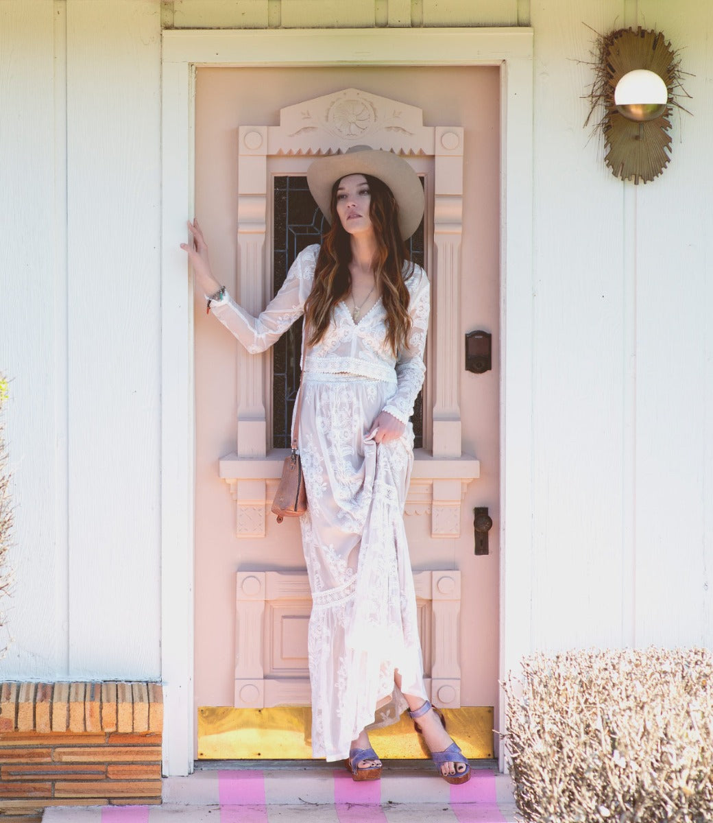 A woman wearing a white dress, cowboy hat and Grettell sandals while standing in front of a pink door.
