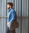 A man wearing a blue shirt and jeans is carrying a Bed Stu Hampton II messenger bag.