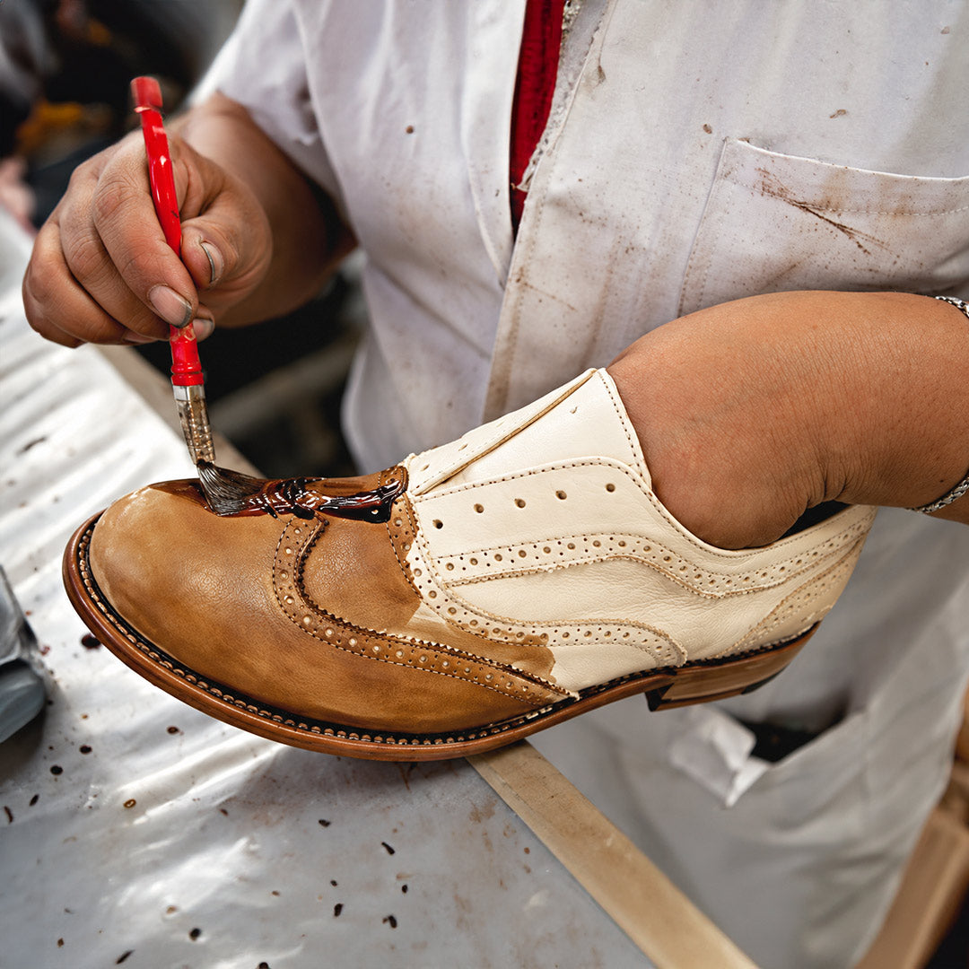 A person in a white lab coat uses a brush to apply dye to a two-tone leather wingtip shoe.