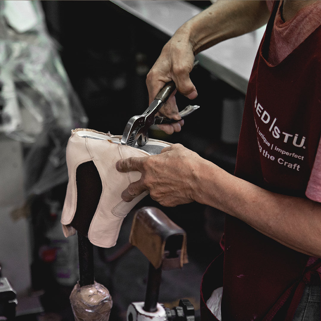 A person wearing a branded apron uses pliers to stretch light-colored leather over a shoe last.