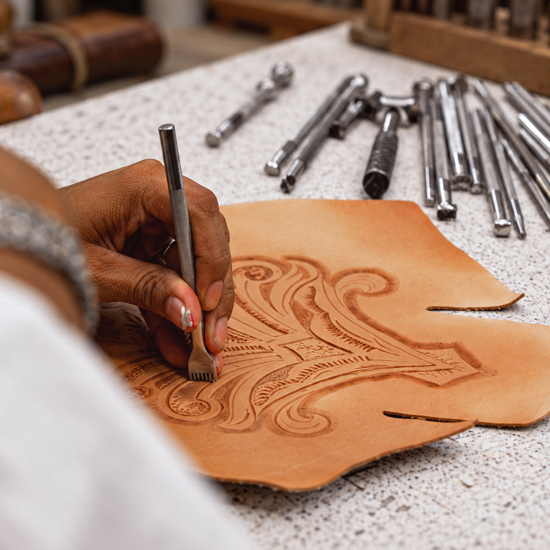 A person uses a metal tool to emboss a decorative pattern into a piece of leather, with various metal stamping tools arranged on the workspace nearby.