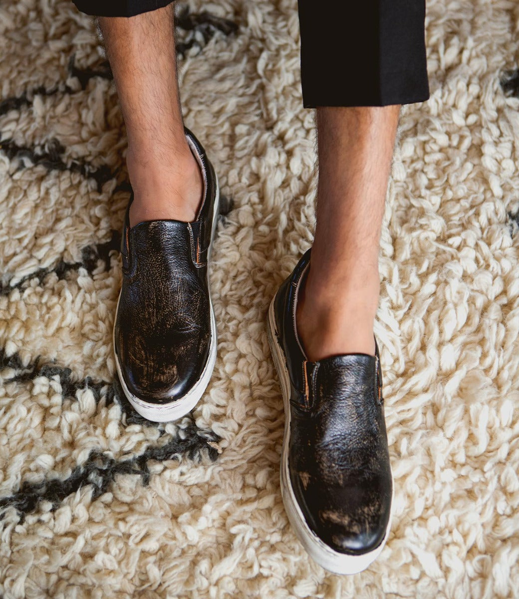 A man wearing a pair of black Harry slip on sneakers by Bed Stu on a rug.